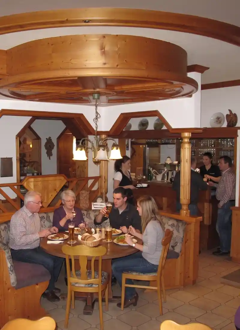 In diesem gemütlichen Gasthaus sitzen vier Gäste an einem Holztisch. Hinter ihnen servieren Mitarbeiter an der Theke Getränke, während andere sich unterhalten. Das rustikale Ambiente mit seinen Holzmöbeln und der warmen Beleuchtung versprüht den klassischen Charme von Ronneburg.